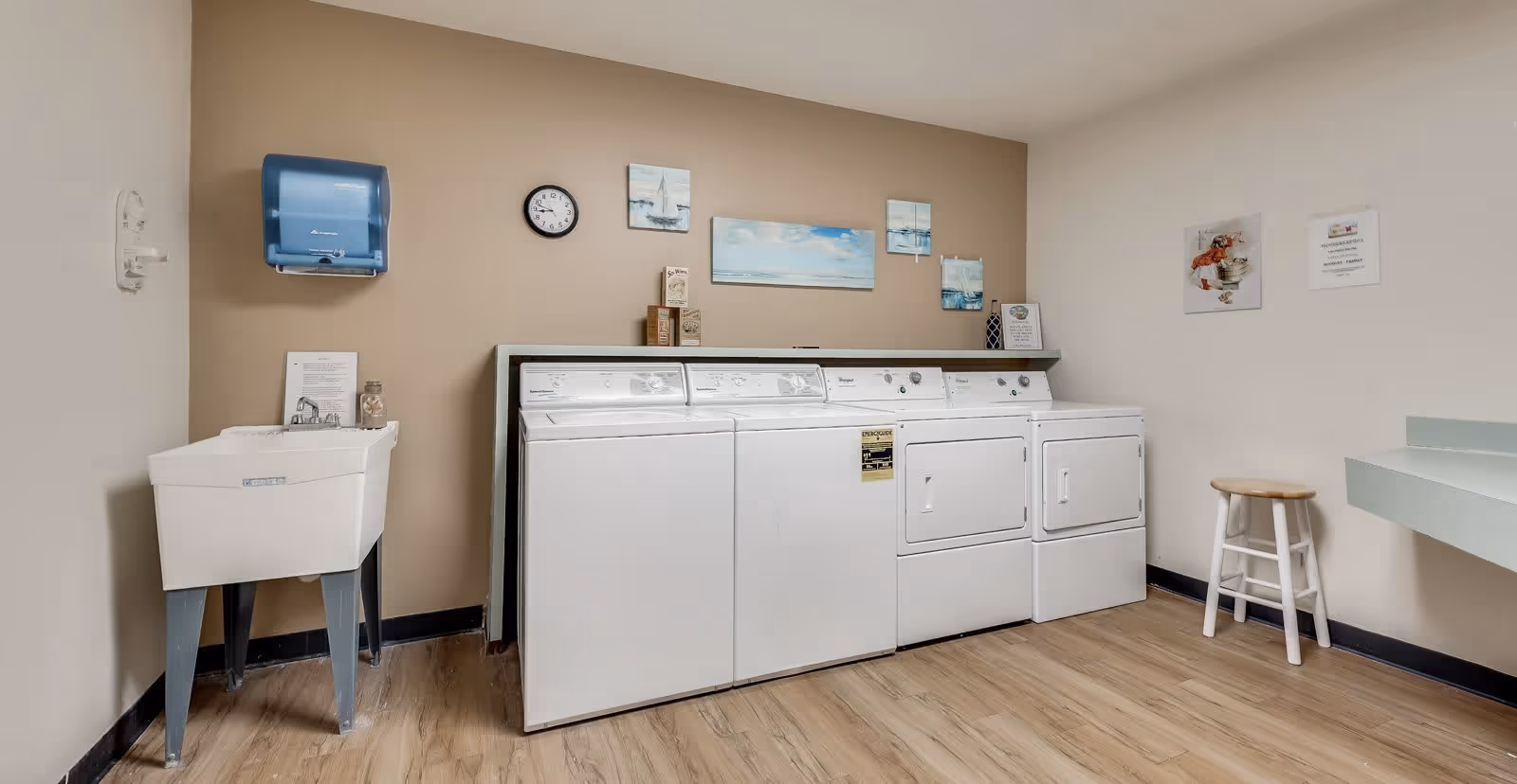 Laundry room with two washing machines and two dryers lined up against a beige wall. Above the machines are several small paintings and a clock. To the left is a utility sink with a blue paper towel dispenser mounted on the wall above it. On the right side, there is a small wooden stool and a countertop. The floor is wood-patterned.