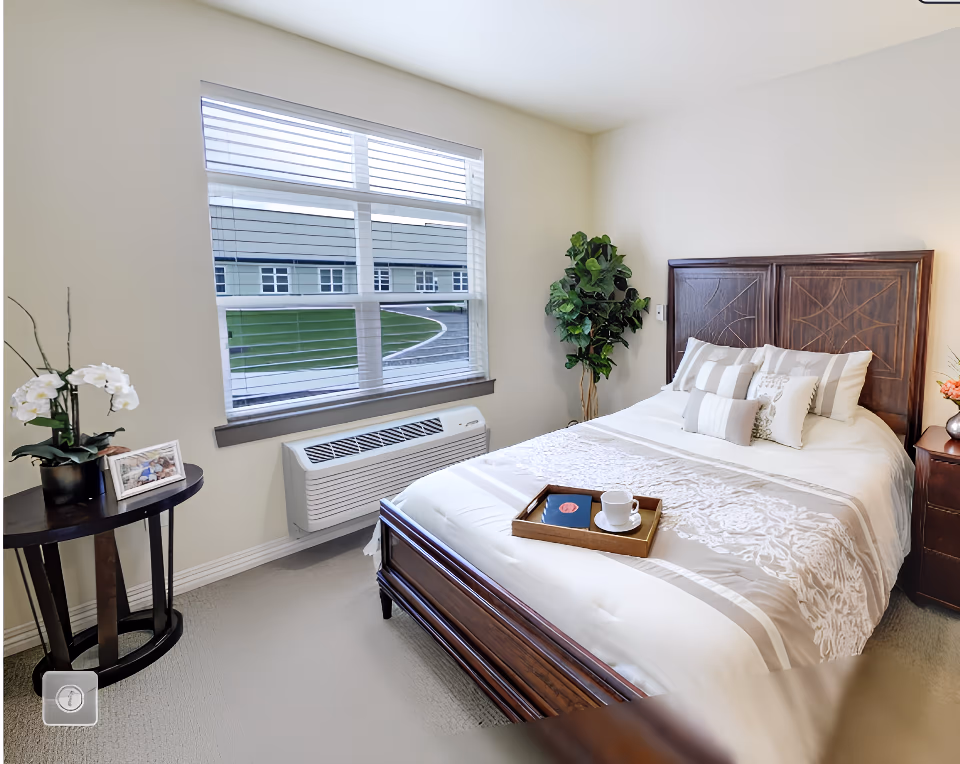 A bright and cozy bedroom with a large window showing an outdoor view. The room features a wooden bed with white and beige bedding, decorative pillows, and a wooden tray holding a cup and a book. There is a small round table with a potted orchid and a framed photo, and a tall green plant in the corner. The walls are light-colored, and there is a heating/cooling unit below the window.
