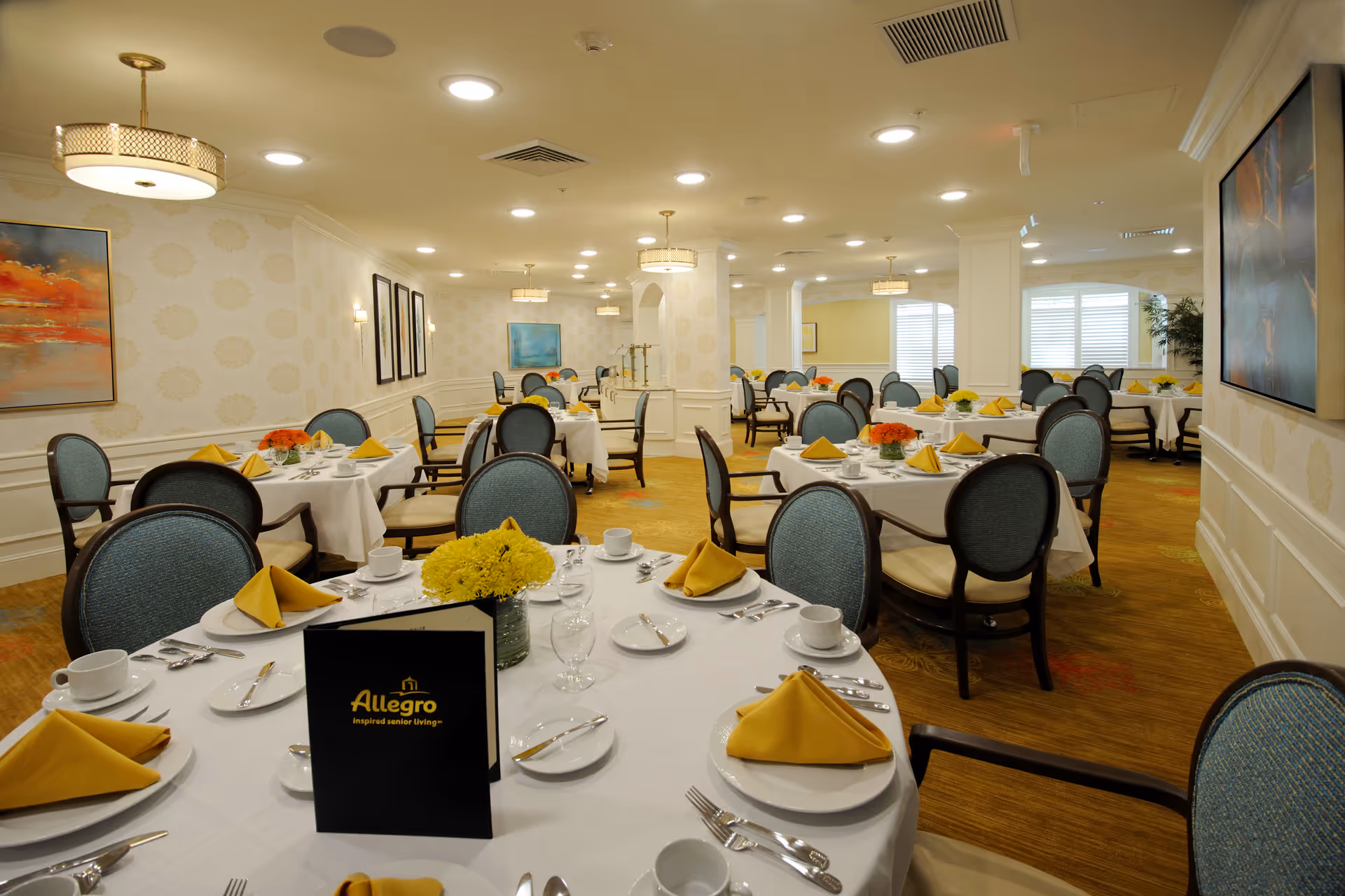 A spacious dining room with multiple round tables covered with white tablecloths, each set with yellow folded napkins, white plates, cups, and silverware. The chairs have blue upholstery and dark wooden frames. The room is well-lit with ceiling lights and decorative hanging lamps. There are paintings on the walls and a menu card on the nearest table displaying the Allegro Senior Living logo.