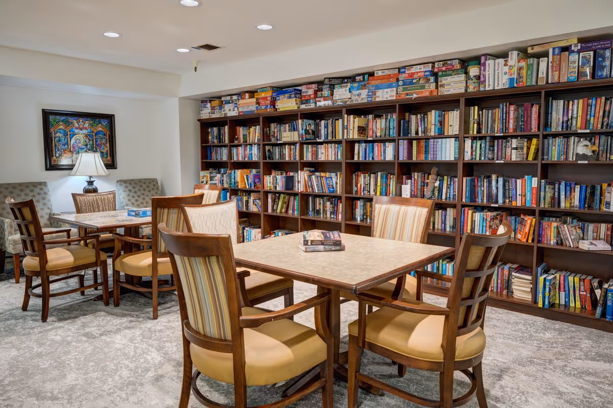A cozy reading room with multiple wooden bookshelves filled with books and board games. There are two square tables with cushioned wooden chairs around them. A lamp and framed artwork are visible on the wall, and the room has a carpeted floor.