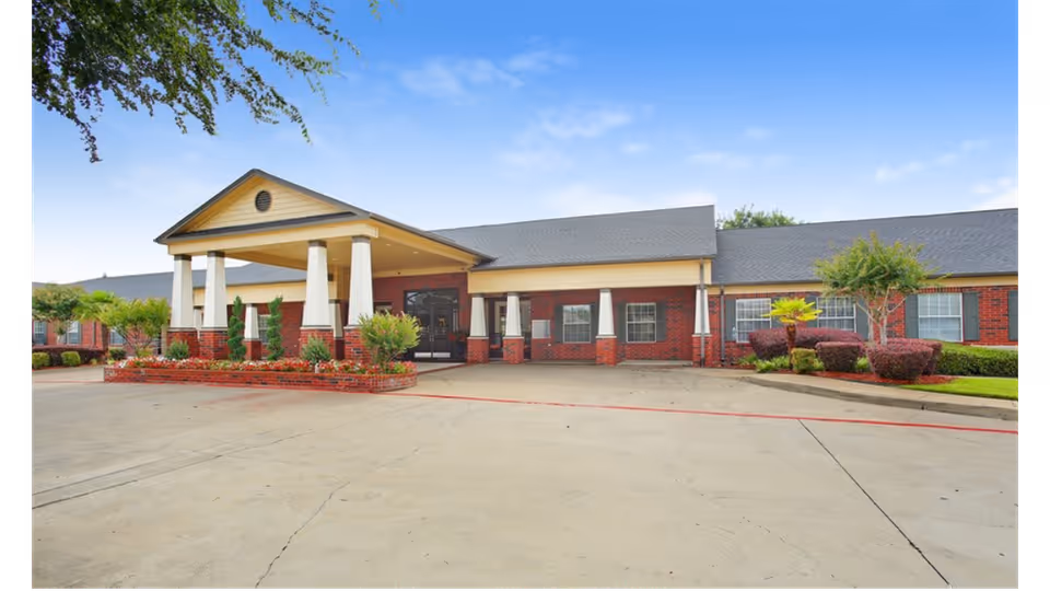Front exterior view of Bedford Estates Senior Living building with a covered entrance supported by white columns, red brick walls, and a paved driveway surrounded by landscaped bushes and trees under a blue sky.