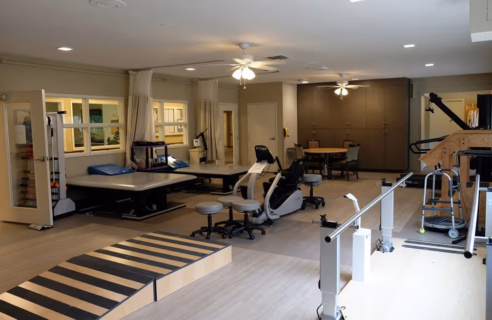 A rehabilitation therapy room with exercise equipment including a recumbent bike, parallel bars for walking practice, therapy tables with blue cushions, stools, and a wooden ramp with black stripes. The room has ceiling fans, beige walls, and a round table with chairs in the background.