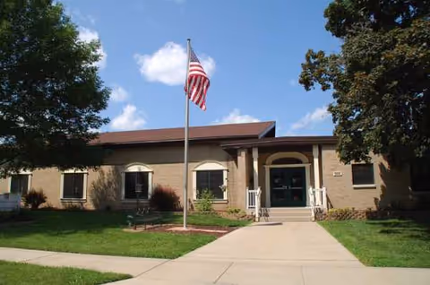 Front view of a single-story assisted living building with flag, entrance stairs, and green lawn.