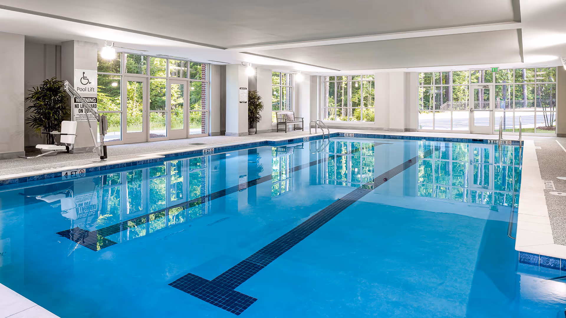 Indoor swimming pool with clear blue water, large windows letting in natural light, pool lift for accessibility, and seating areas along the walls.