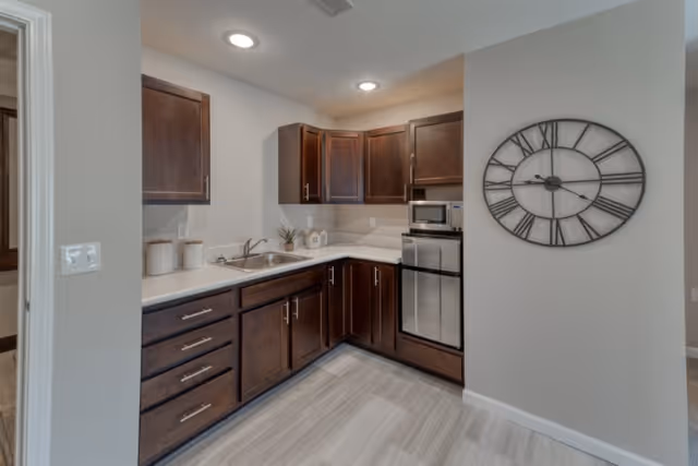 A modern kitchen with dark wooden cabinets, white countertops, a stainless steel mini refrigerator, and a microwave. The kitchen has a sink with a faucet, three canisters on the counter, and a small plant decoration. There is a large decorative wall clock with Roman numerals on the adjacent wall. The floor has light-colored tiles, and the walls are painted light gray.