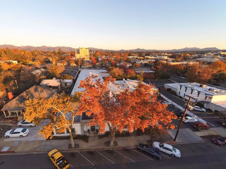 Aerial view of a neighborhood with autumn-colored trees and buildings, including a white flat-roofed building in the center, several parked cars along the street, and mountains in the background under a clear sky.
