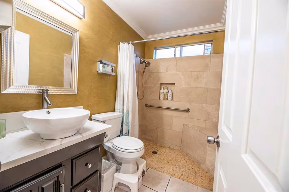 A bathroom with a white vessel sink on a marble countertop, a large framed mirror above the sink, a toilet with a small step stool in front, and a walk-in tiled shower with a built-in bench, grab bar, and shower curtain. The walls are painted mustard yellow and there is a window above the shower.