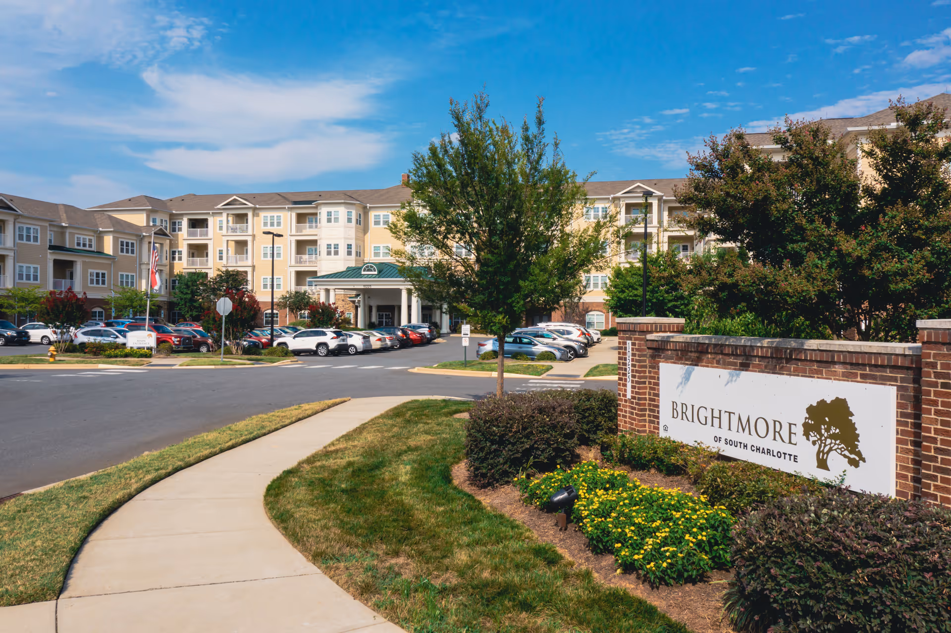 Exterior view of Brightmore of South Charlotte senior living facility showing a multi-story building with balconies, a parking lot with cars, a sidewalk, landscaped greenery, and a sign with the facility's name.