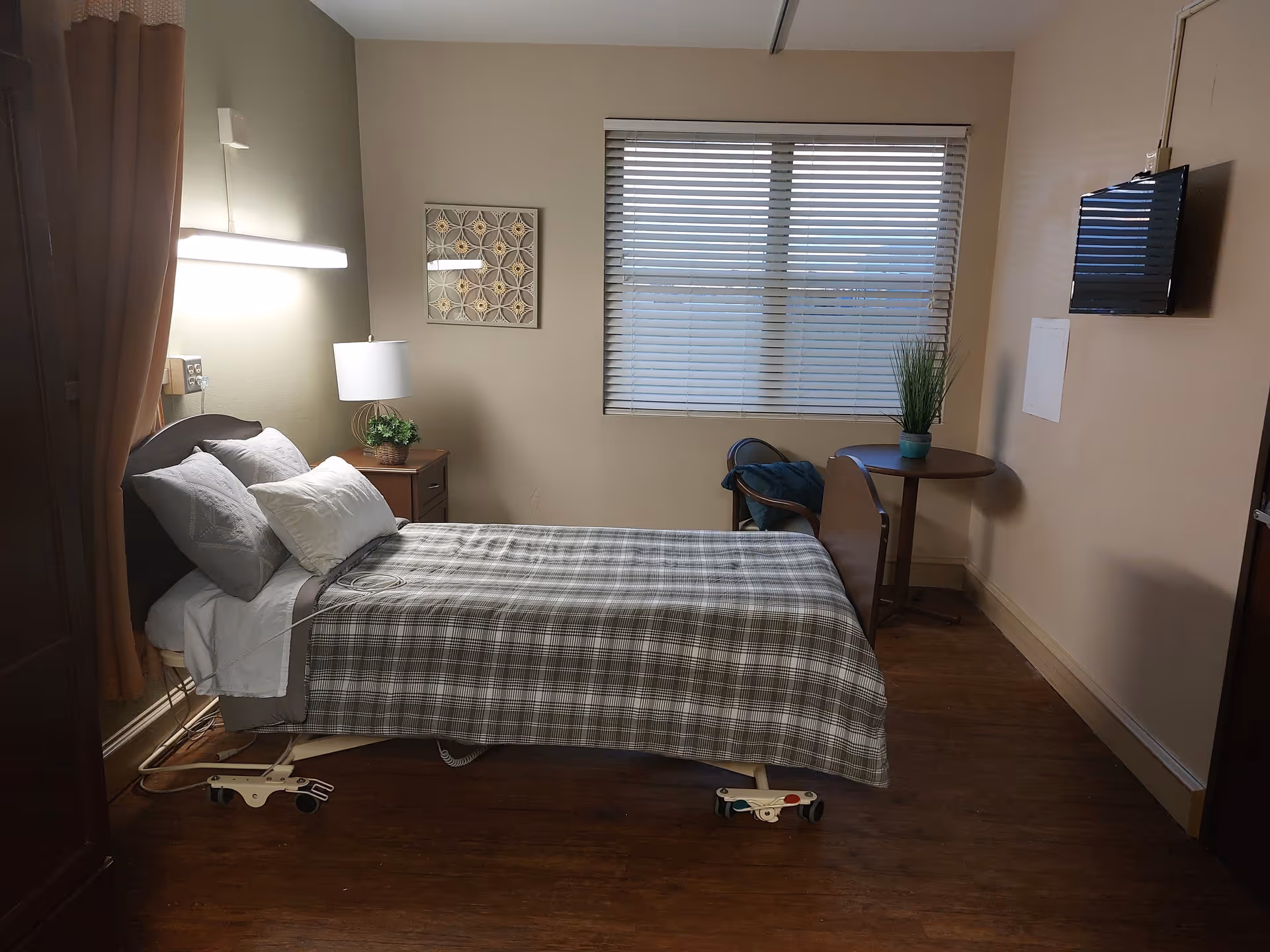A tidy nursing facility bedroom with a single hospital-style bed, bedside table and lamp, window with blinds, small round table and wall-mounted TV.