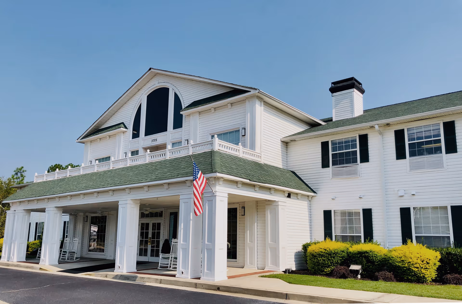 Exterior view of a two-story white senior living facility building with green roof shingles, large windows, a covered entrance with white columns, rocking chairs on the porch, an American flag, and well-maintained landscaping with bushes and grass under a clear blue sky.
