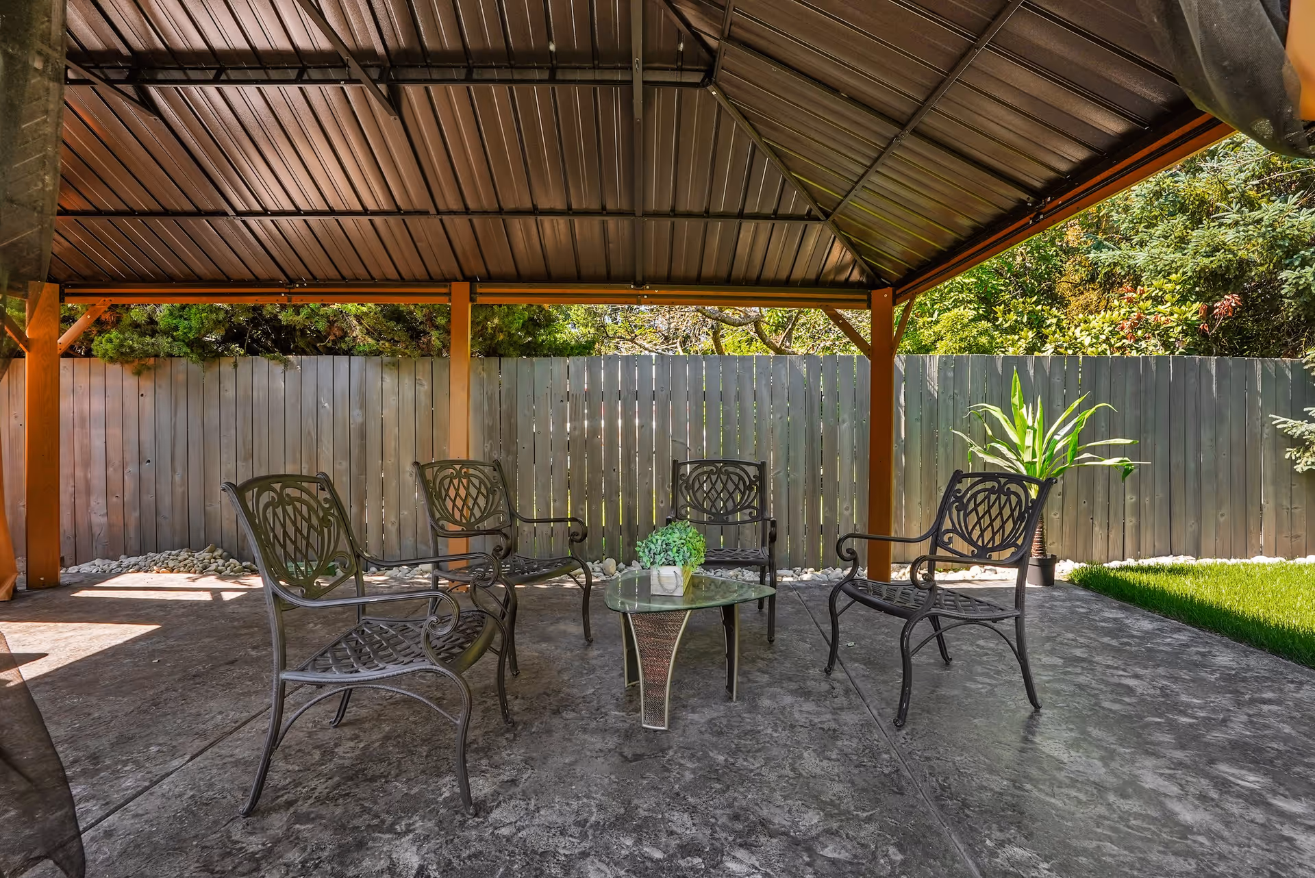 Covered outdoor patio area with four metal chairs arranged around a small glass-top table with a potted plant, surrounded by a wooden fence and greenery.