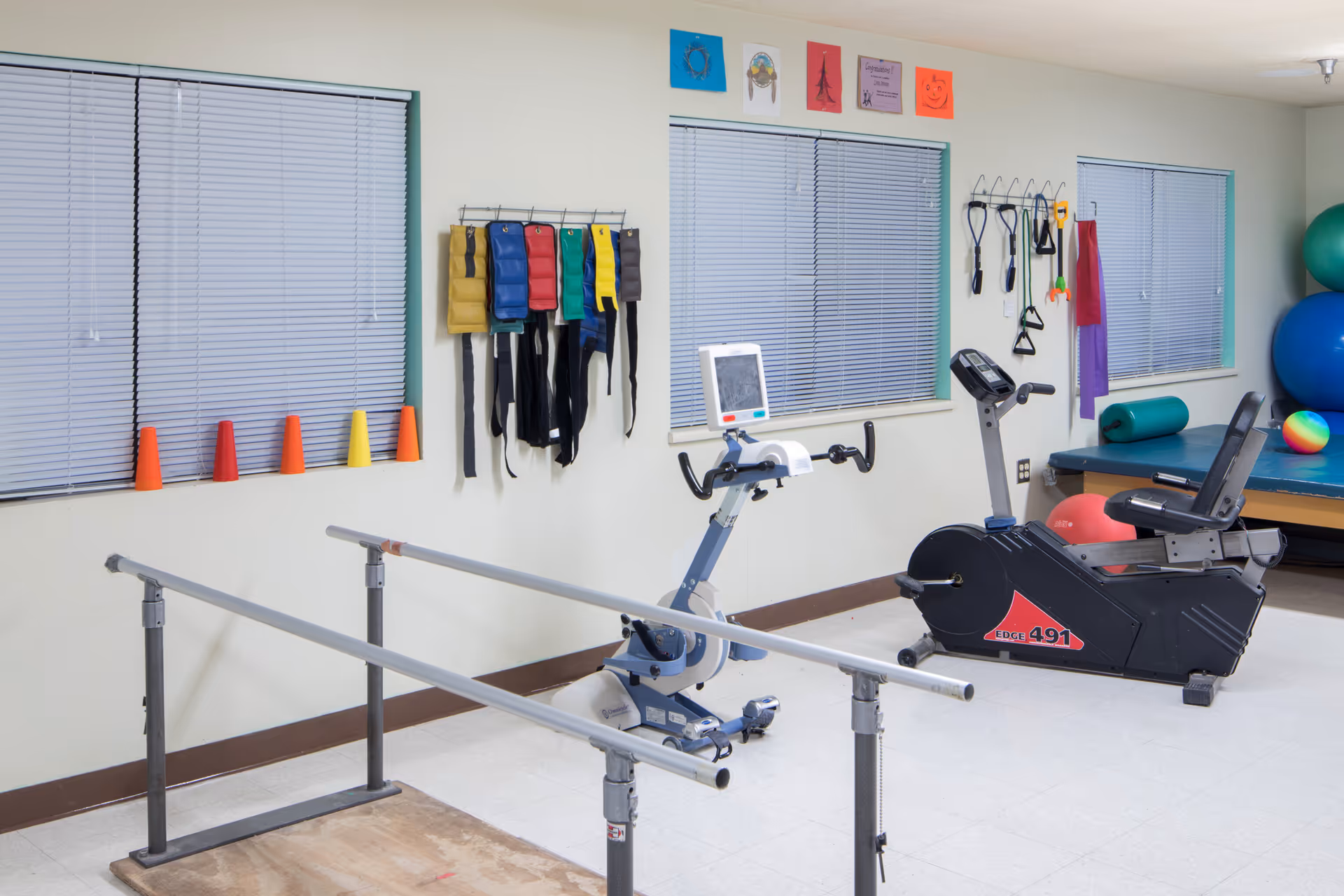 A physical therapy room with parallel bars for walking exercises, a stationary recumbent bike, various exercise equipment hanging on the wall, colorful cones on the windowsill, and large exercise balls in the corner.