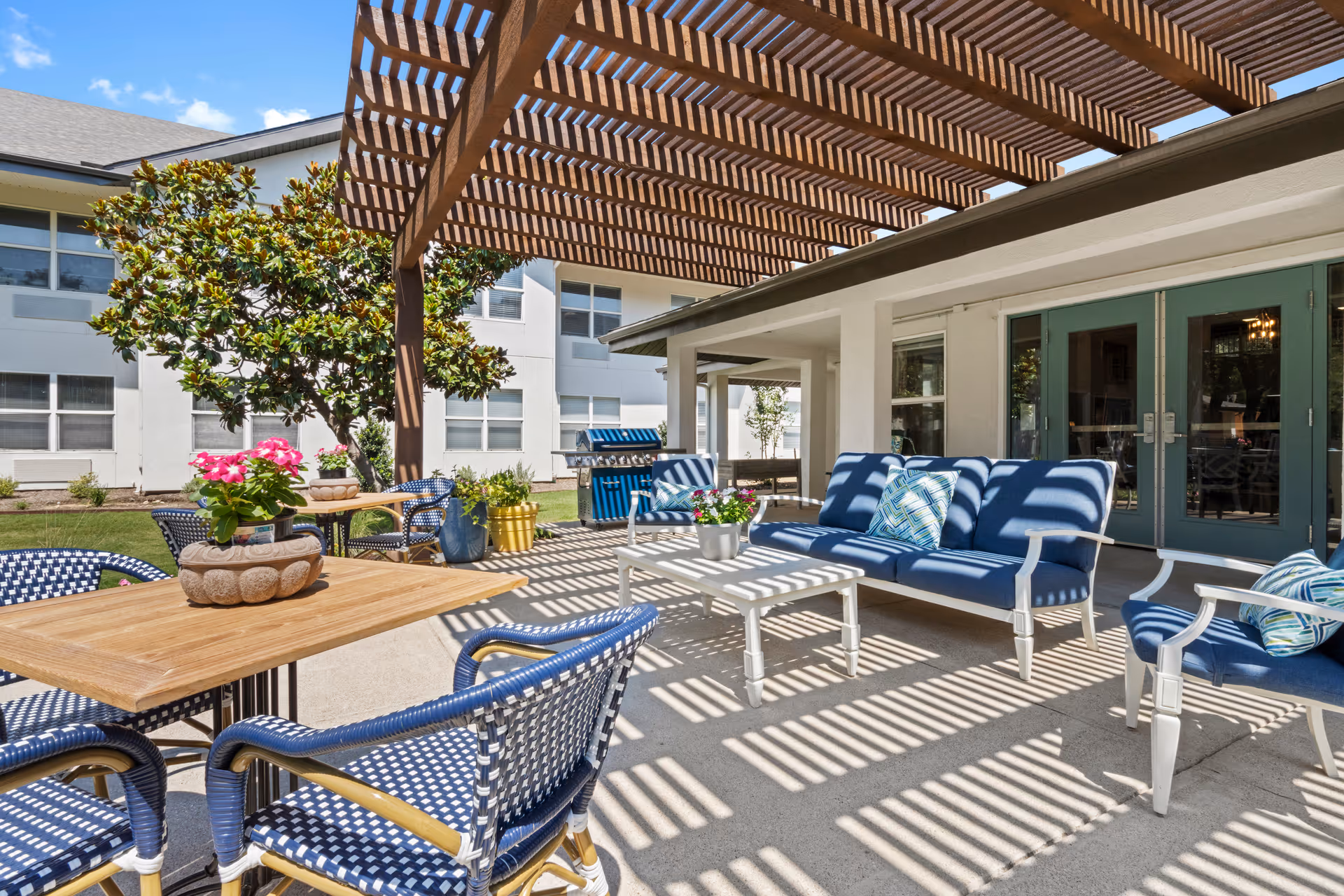 Outdoor patio with a wooden pergola casting striped shadows over blue cushioned seating, tables, and potted plants.