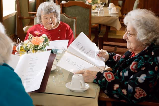 Three elderly women sitting around a dining table in a restaurant or dining room, looking at menus. The table has a floral centerpiece, a cup and saucer, and some condiments. The background shows other tables with tablecloths and chairs.