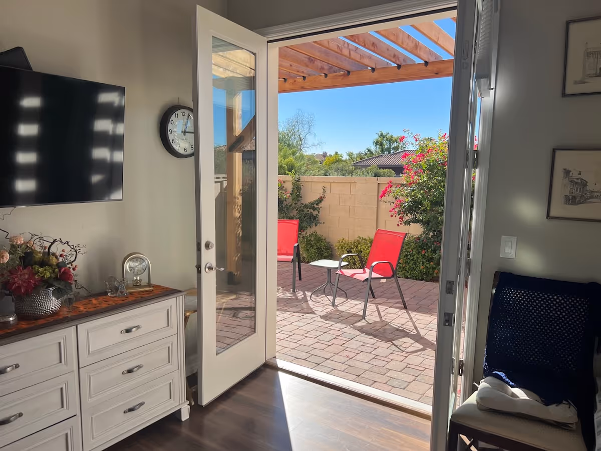 View from inside a living room looking out through an open glass door to a sunny patio area with two red chairs and a small table under a wooden pergola. Inside, there is a white dresser with a flower arrangement and a clock on it, a wall-mounted TV, a wall clock, and a chair with a dark blue knitted blanket.