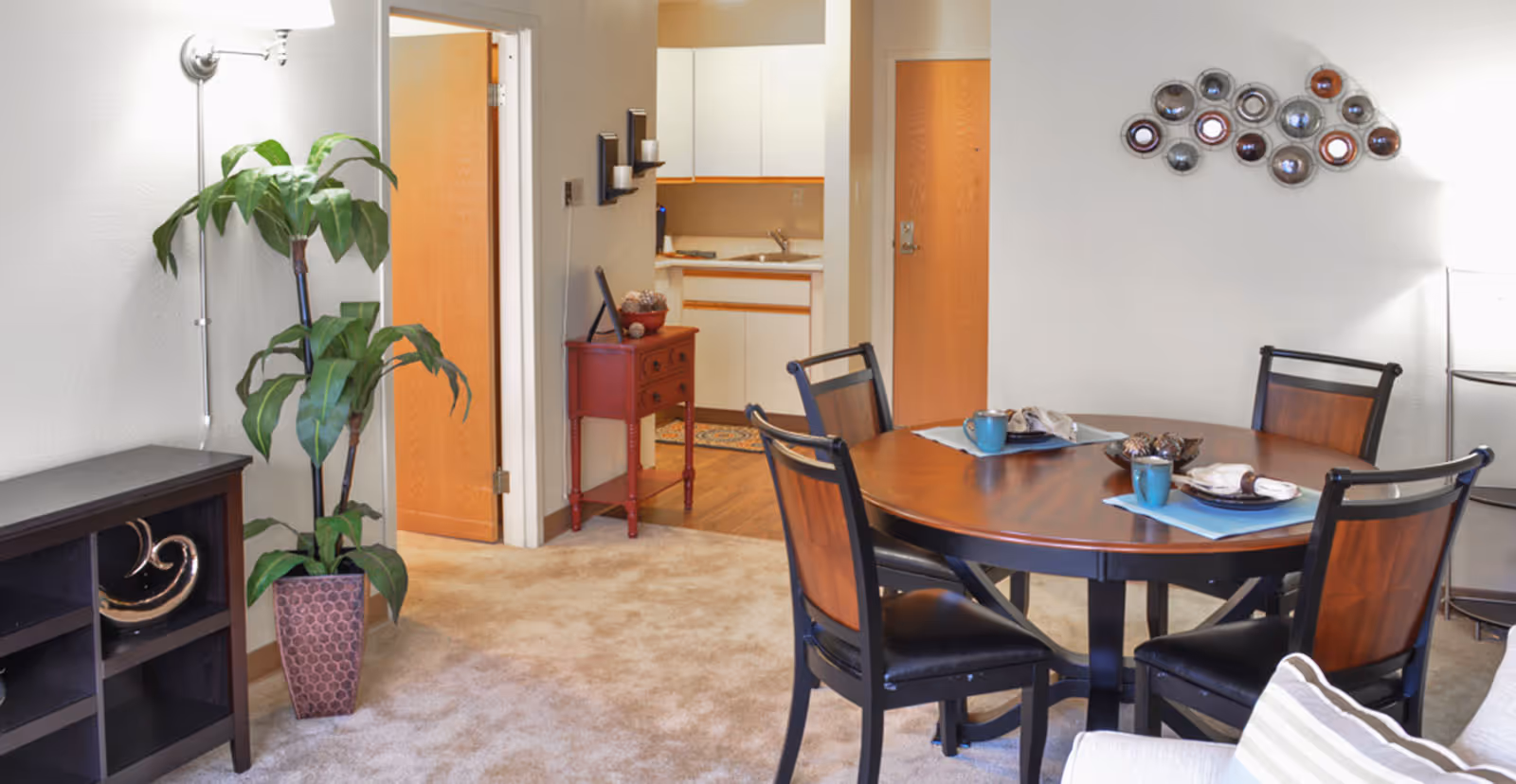 A cozy interior dining area with a round wooden table set for four, chairs, a potted plant, and a view into a small kitchenette.