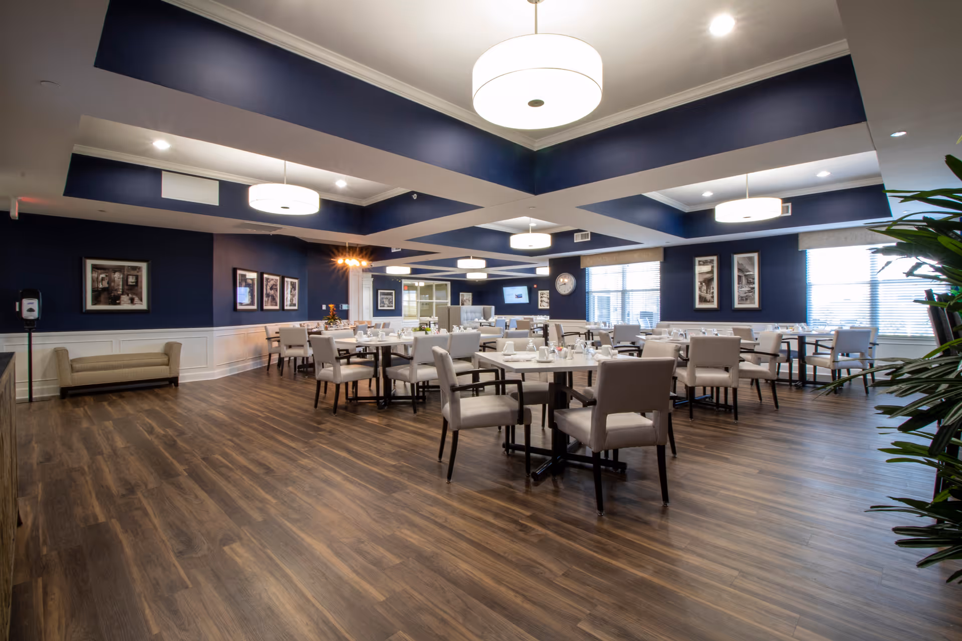 A spacious dining room with multiple tables and chairs arranged neatly. The room features dark blue walls with white wainscoting, large windows with blinds, modern ceiling lights, and wood flooring. There are framed pictures on the walls and a clock near the windows.