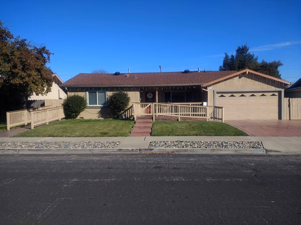 Single-story residential building with a brown roof and beige exterior walls. There is a wheelchair accessible ramp leading to the front door, flanked by bushes and a small lawn. The house has a two-car garage on the right side and a clear blue sky overhead.
