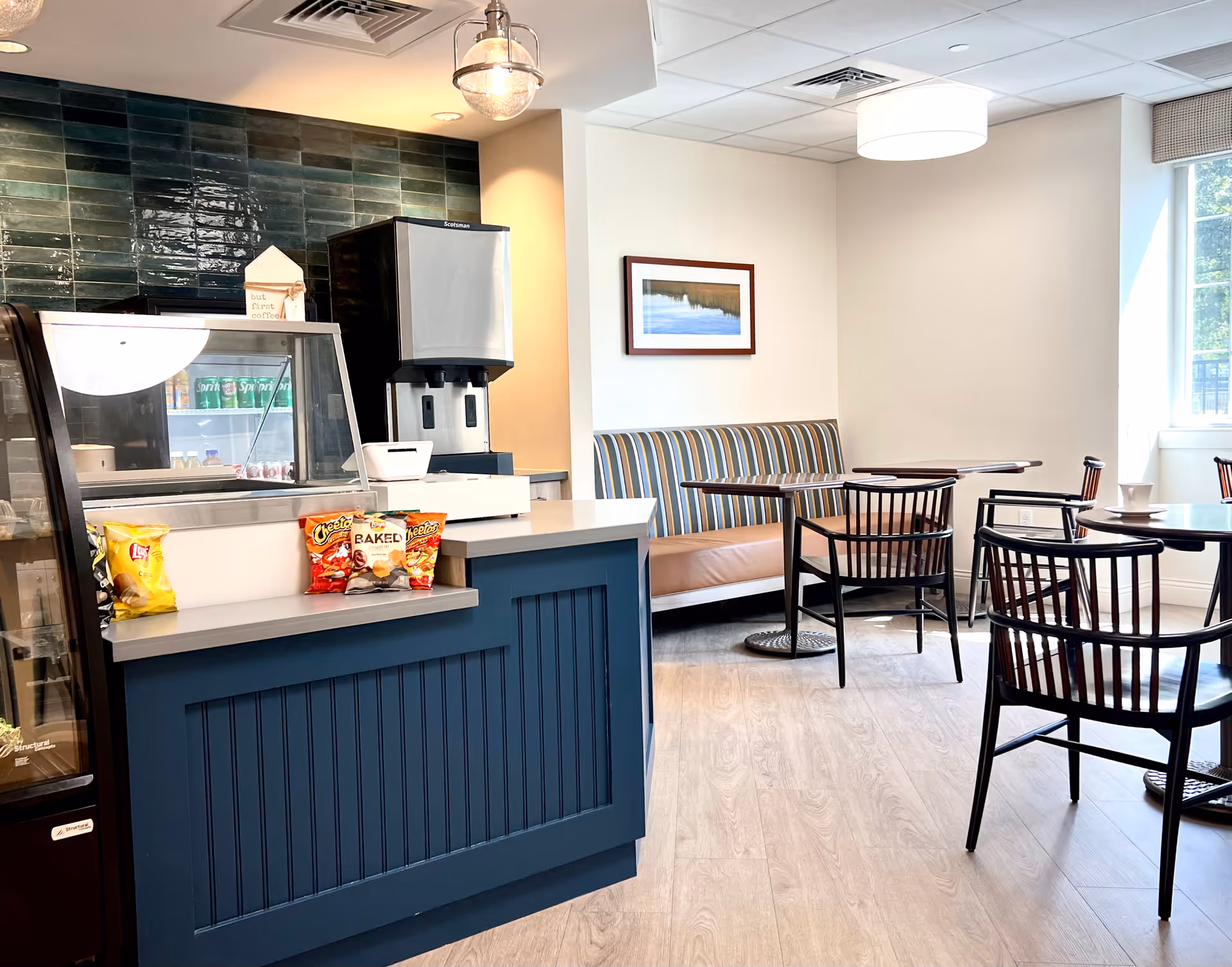 A small dining area with a blue counter displaying snack bags including Lay's and Cheetos. Behind the counter is a beverage dispenser and a refrigerated display case with drinks. The seating area has wooden chairs and tables, with a striped cushioned bench against the wall and a framed landscape picture above it. Large windows allow natural light into the room.