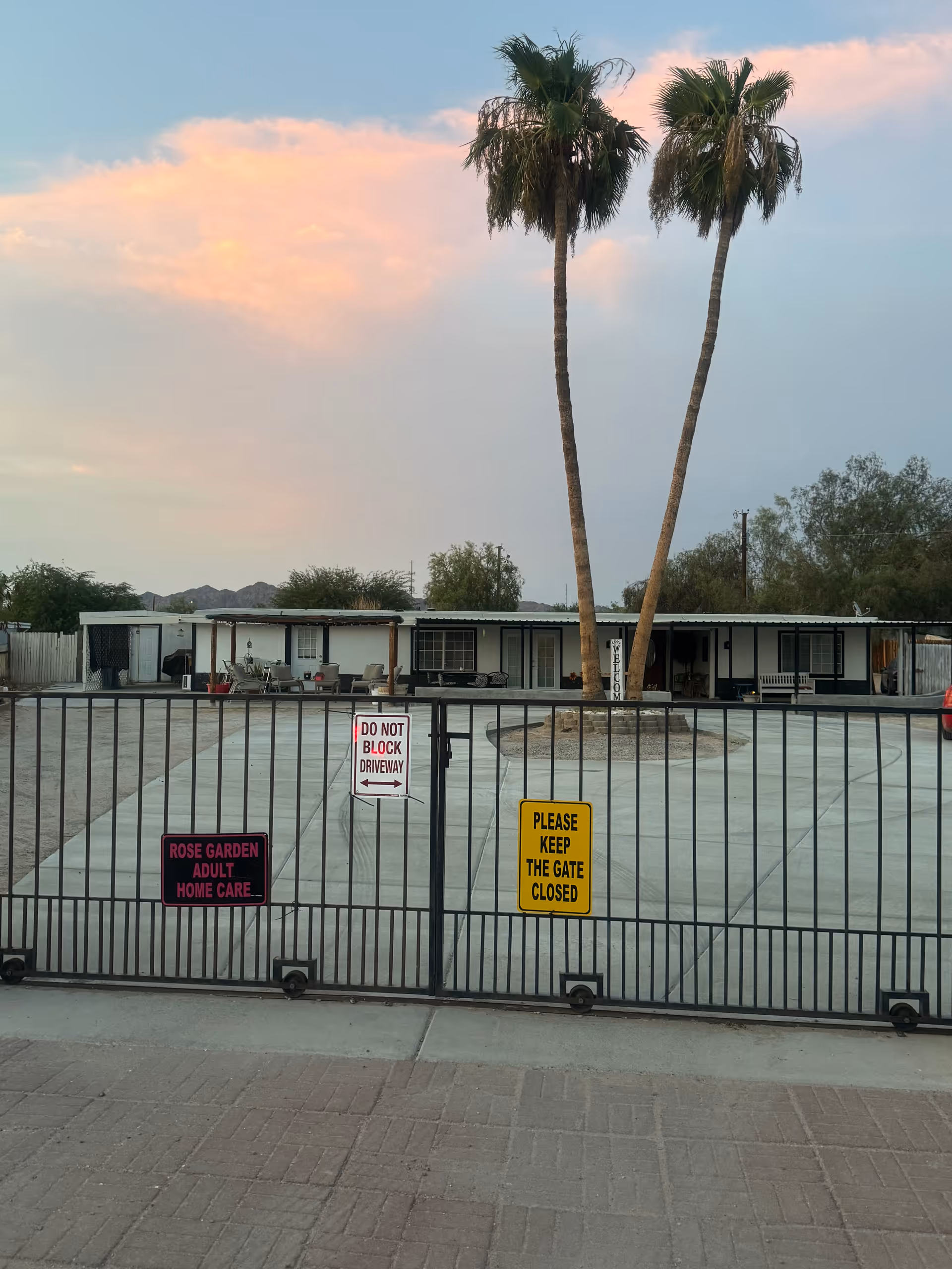 View of the front entrance of Rose Garden Assisted Living, showing a gated driveway with signs that read 'ROSE GARDEN ADULT HOME CARE,' 'DO NOT BLOCK DRIVEWAY,' and 'PLEASE KEEP THE GATE CLOSED.' Behind the gate is a single-story building with a covered porch, outdoor seating, and two tall palm trees. The sky is partly cloudy with a soft sunset glow.