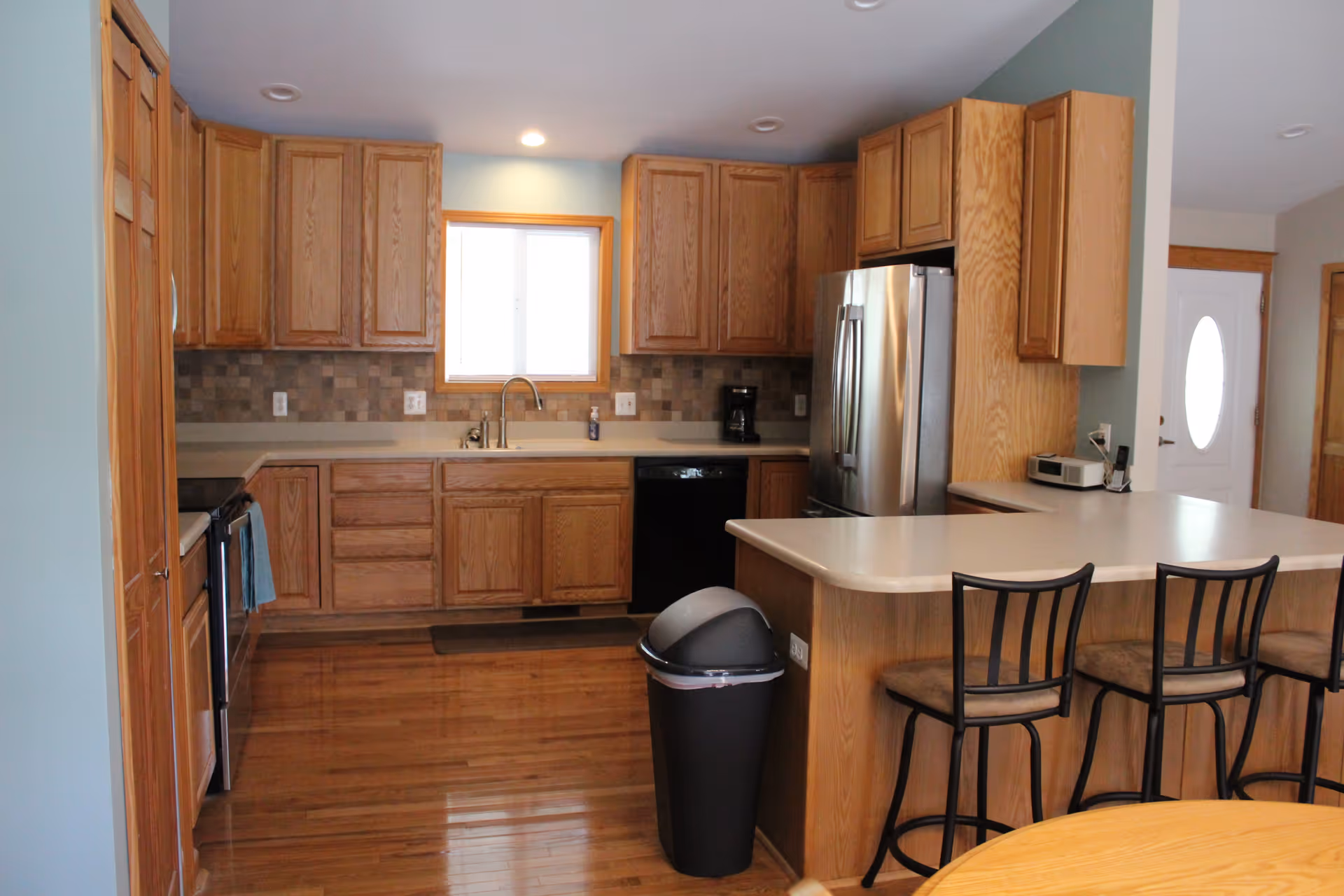 Open kitchen with wooden cabinets, a center island with bar stools, stainless steel refrigerator, sink under a window, and a trash can on hardwood floors.