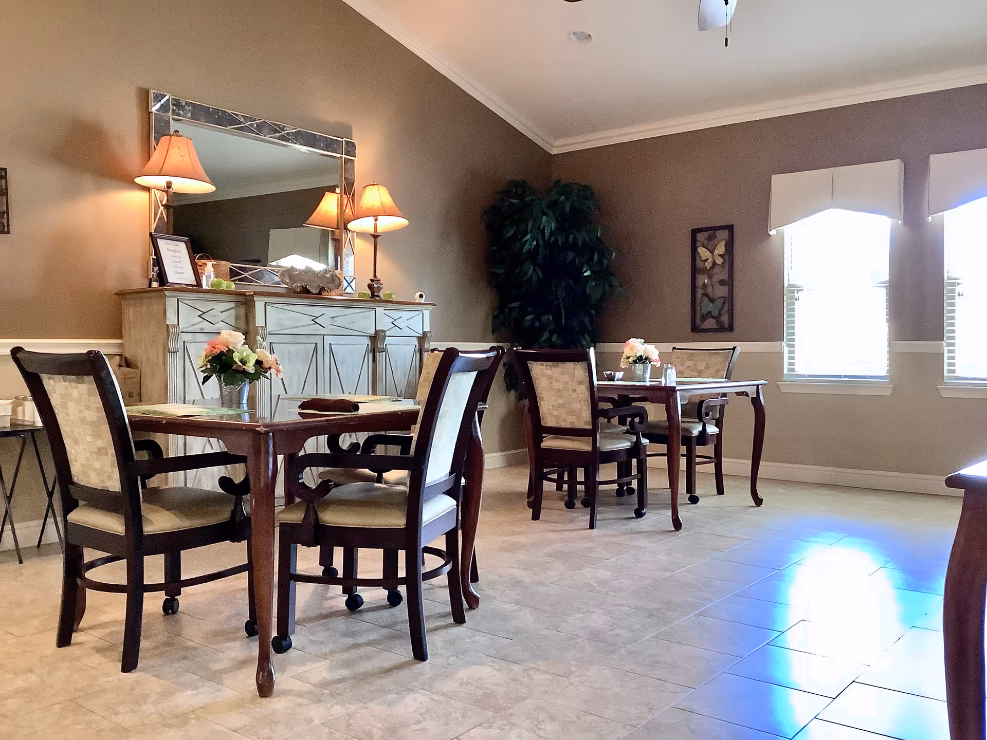 Dining room with wooden tables and upholstered chairs, a decorative sideboard with lamps and mirror, and windows letting in natural light.