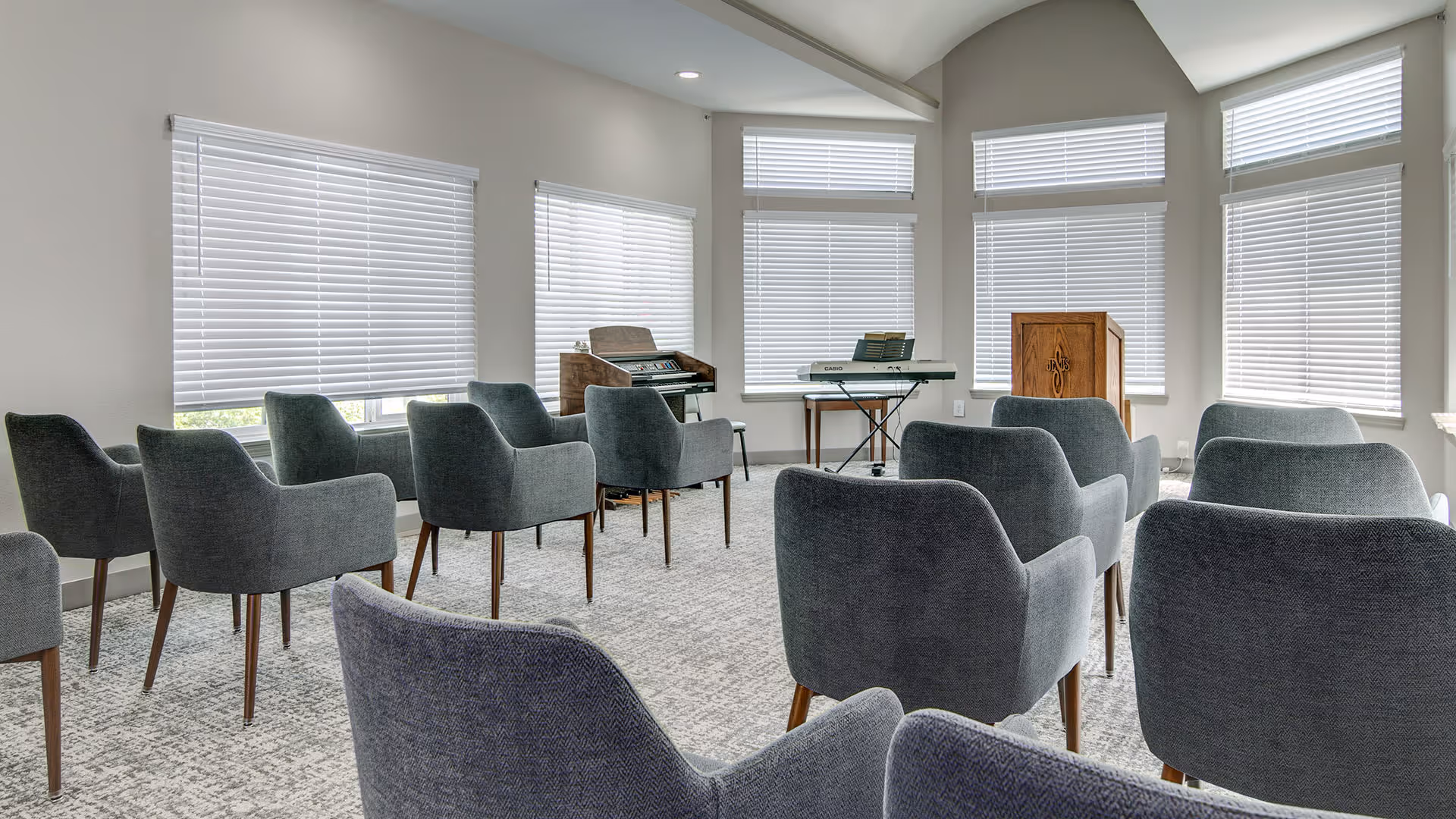 A bright room with multiple gray upholstered chairs arranged in rows facing a wooden podium and two electronic keyboards near windows with white blinds.