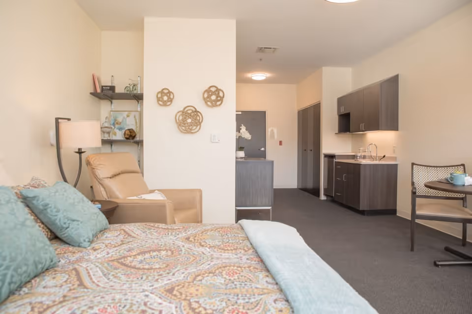 A cozy senior living facility room featuring a bed with patterned bedding and teal pillows, a tan recliner chair beside a floor lamp, a small round table with a chair and a cup, and a kitchenette with dark wood cabinets and a sink. The room has neutral walls and carpeted flooring, with decorative wall art and shelving.