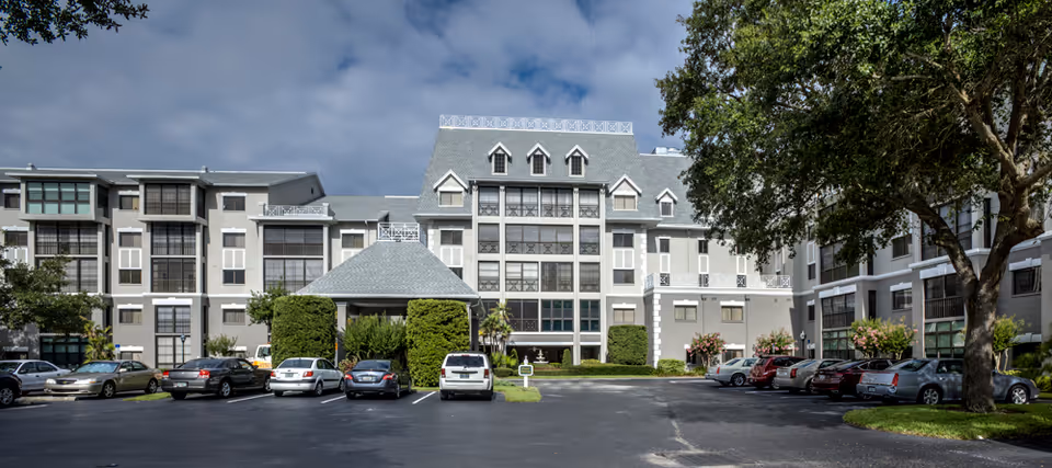 Front exterior of a multi-story senior living building with a driveway, parked cars, and trees under a partly cloudy sky.