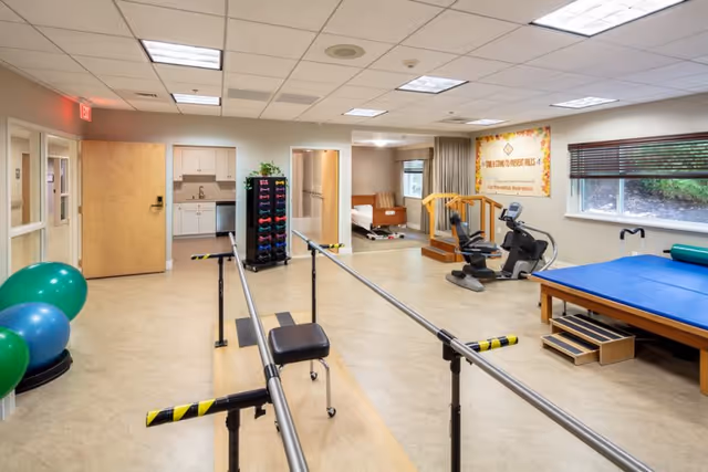 A rehabilitation room in Belmont Manor Nursing & Rehabilitation Center featuring parallel bars for walking exercises, exercise balls, a rack of dumbbells, a recumbent exercise bike, a padded therapy table with steps, and a hospital bed in a small adjacent area. The room has beige flooring, white walls, fluorescent ceiling lights, a large window with blinds, and a motivational poster on the wall.