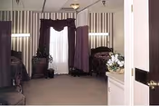 Interior view of a bedroom with two beds separated by a curtain, a window with sheer and dark curtains, a small wooden nightstand with a plant, and a carpeted floor.