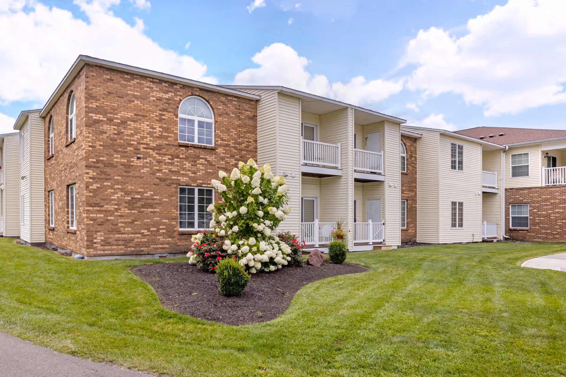 Exterior view of a two-story senior living facility building with a combination of brick and beige siding. The building features multiple windows and small balconies. In front of the building, there is a well-maintained lawn with a landscaped garden area containing white and red flowering bushes. The sky is partly cloudy.