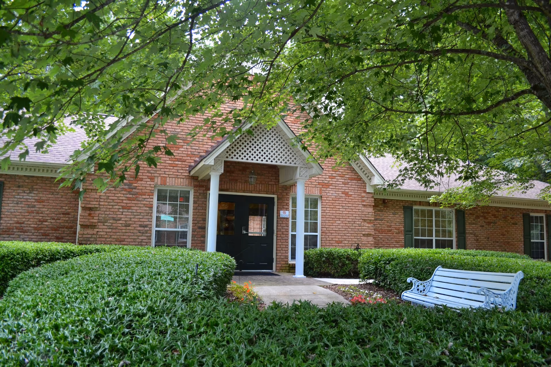Front entrance of a single-story brick building with white columns, trimmed hedges, and a white bench under trees.