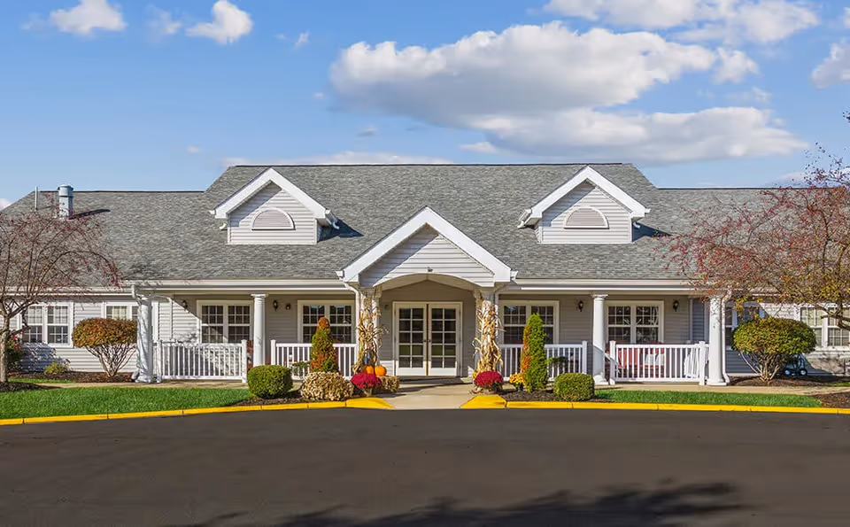 Front exterior view of a single-story senior living facility building with a gray roof, white siding, and a covered entrance. The entrance is decorated with autumn-themed plants and pumpkins. There are small bushes and trees on either side of the entrance, and a clear blue sky with some clouds above.