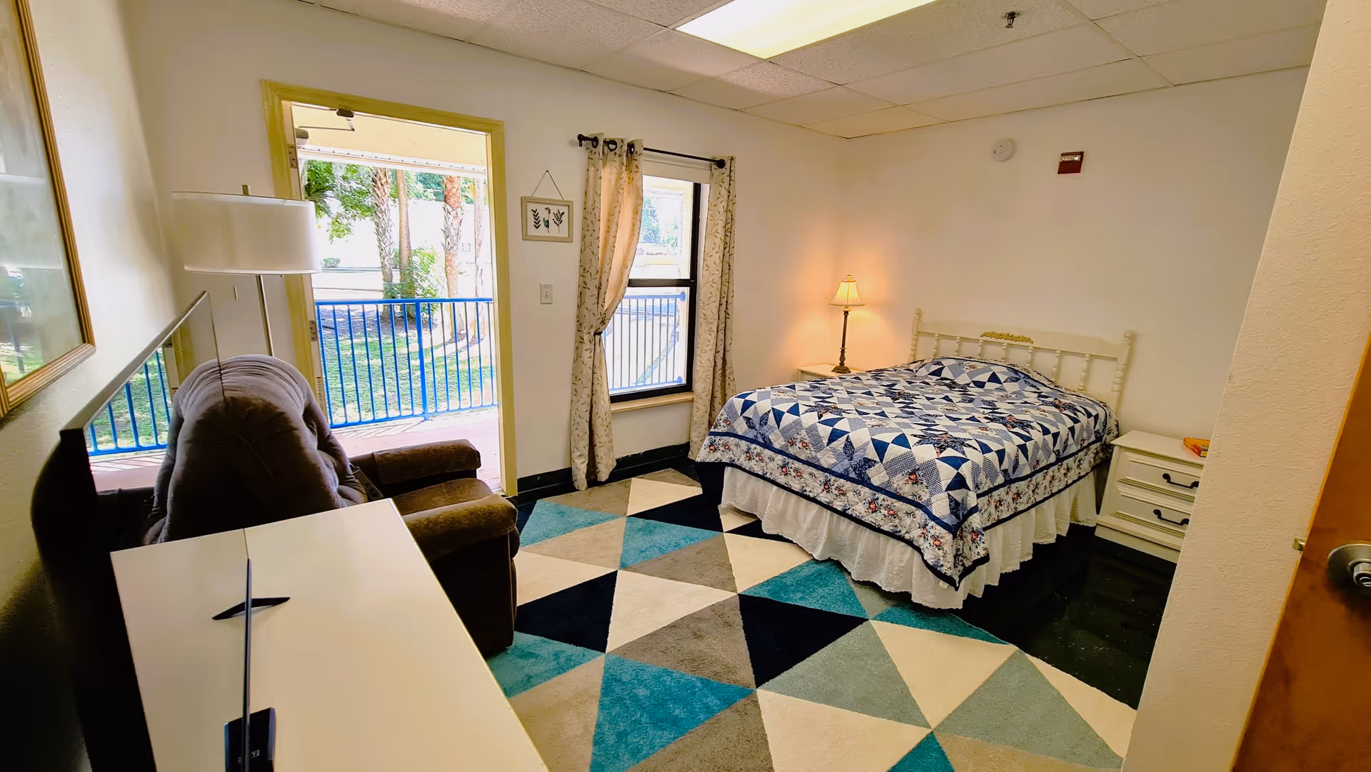 A cozy bedroom with a bed covered in a blue and white patterned quilt, a bedside table with a lamp, a recliner chair, a TV on a white stand, a colorful geometric patterned rug, a window with curtains, and a door leading to an outdoor balcony with a view of trees and grass.