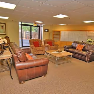 A cozy senior living community lounge area with two brown leather sofas and two cushioned armchairs arranged around a wooden coffee table. The room has beige walls, a carpeted floor, ceiling lights, and a large glass door leading outside. There are decorative pillows on the sofas and a flower arrangement on a side table.