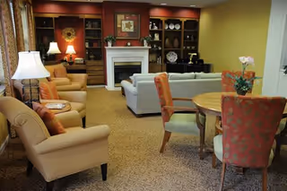 A cozy senior living room with beige armchairs, a white sofa, a round wooden table with four upholstered chairs, a fireplace with a mantel, built-in wooden shelves with decorative items, and warm lighting from table lamps.
