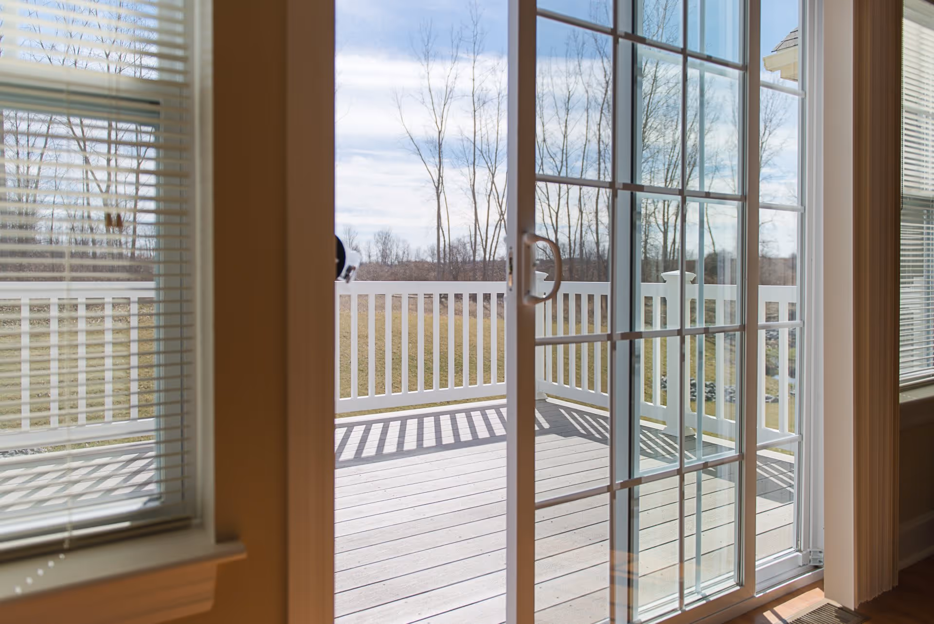 View through a sliding glass door with white grid patterns leading to a wooden deck with white railing. Outside, leafless trees and a grassy area are visible under a partly cloudy sky.