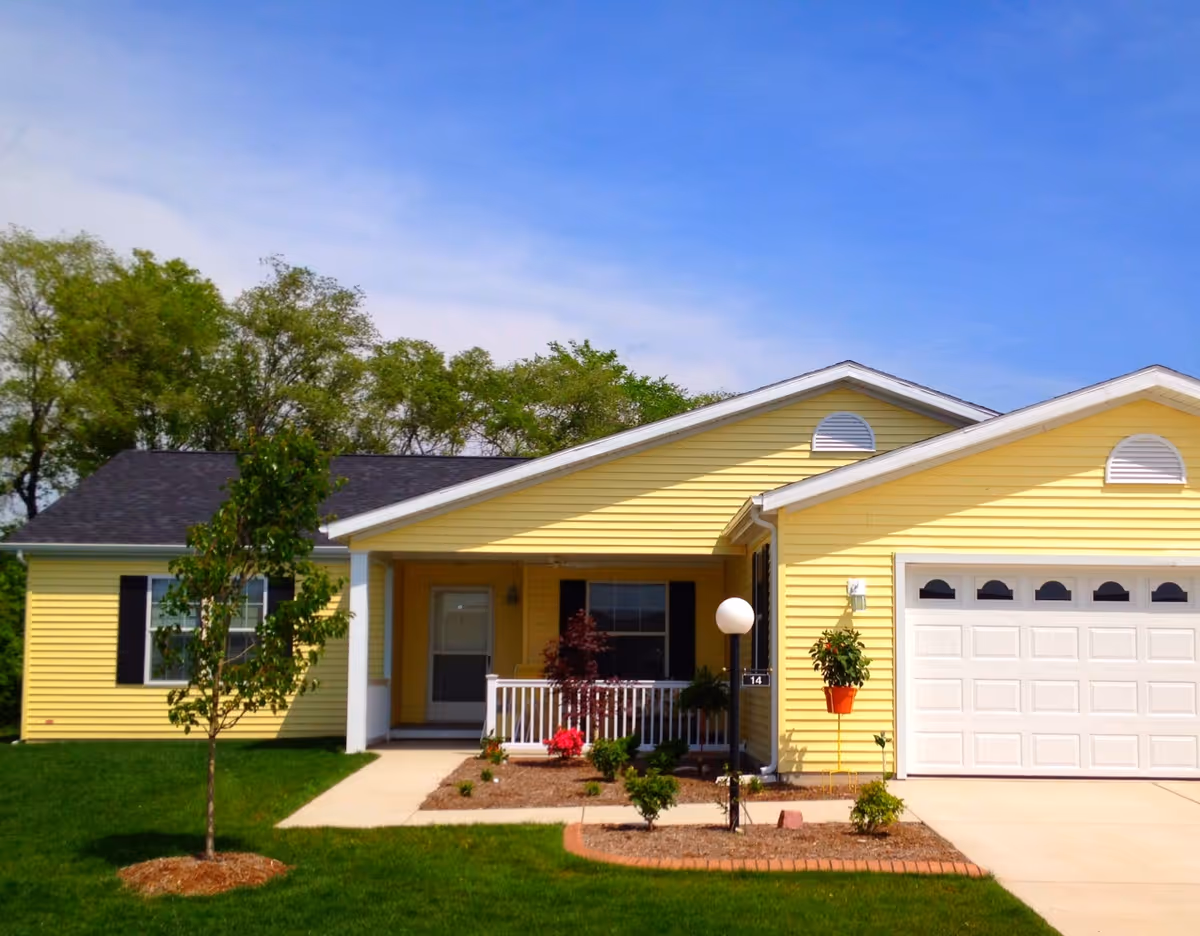 A single-story yellow house with white trim and a black roof. The house has a small front porch with white railings, a white front door, and a large white garage door. There are small shrubs and plants in the front garden, a young tree on the lawn, and a clear blue sky in the background.