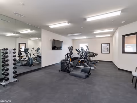 A fitness room with exercise equipment including stationary bikes and treadmills. There is a large mirror on one wall, a wall-mounted TV, and a framed motivational poster. The room has white walls, black flooring, and fluorescent ceiling lights.