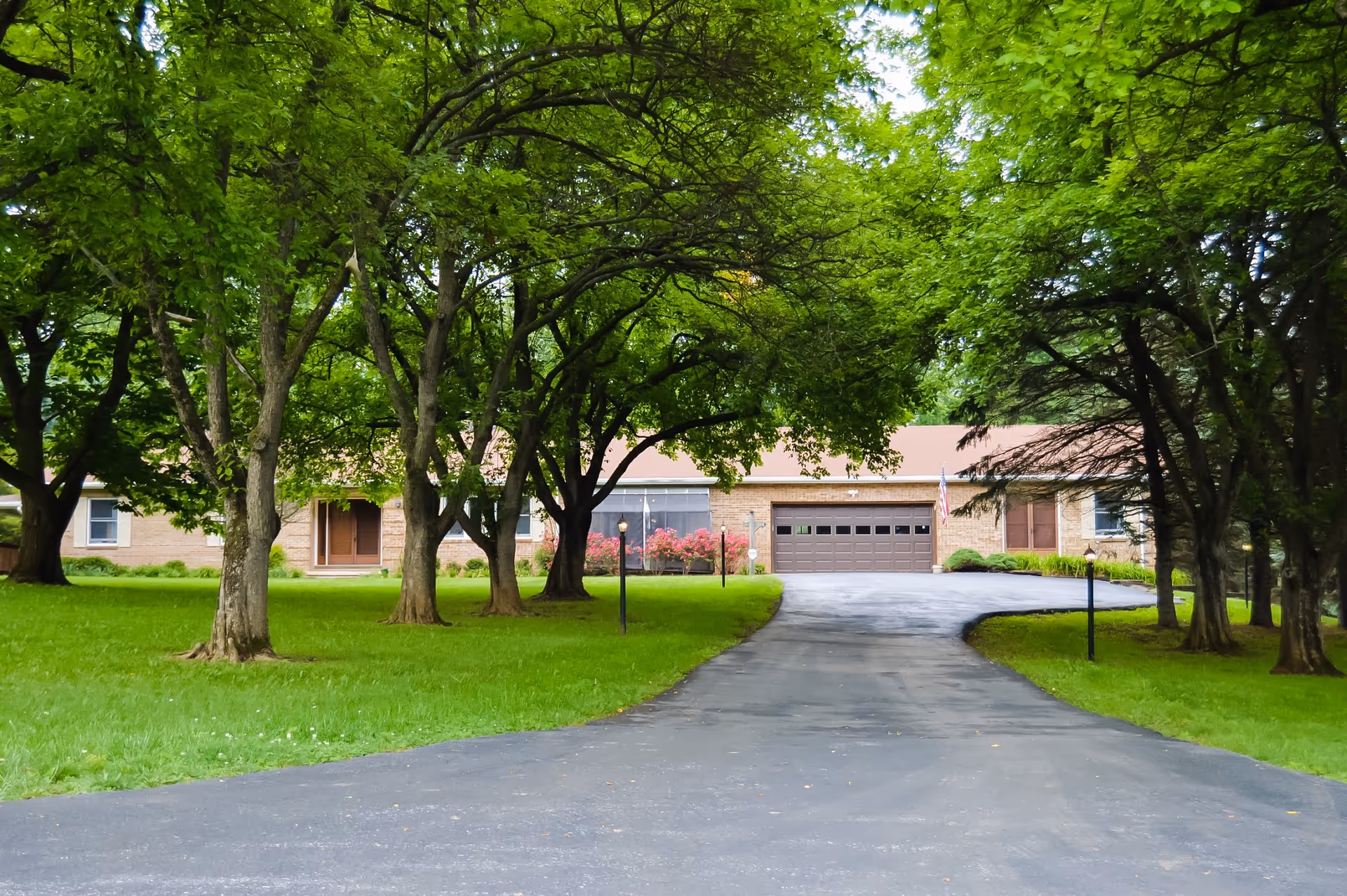Driveway lined with trees leading to a single-story brick building with a garage and manicured lawn.