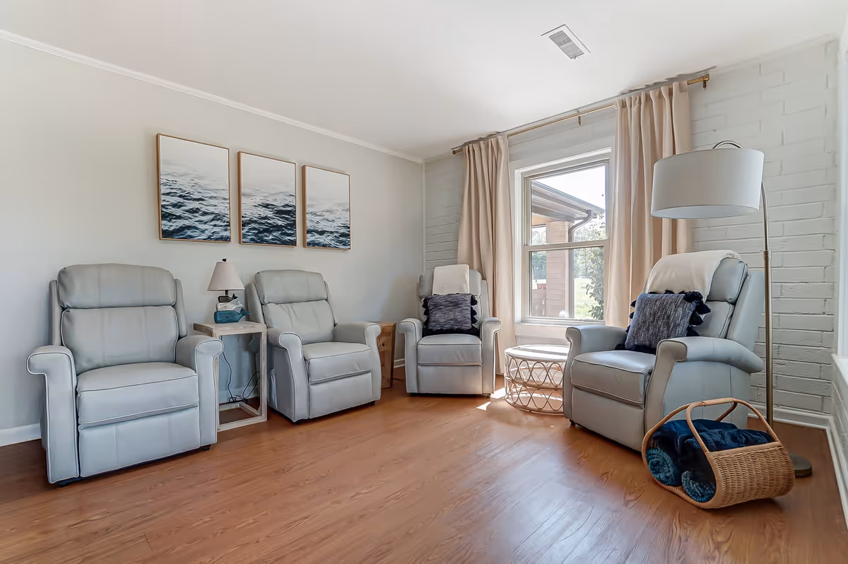 A bright and cozy living room with four light gray recliner chairs arranged around a window with beige curtains. There is a small side table with a lamp between two chairs, a round wicker coffee table near the window, and a floor lamp next to one chair with a basket holding folded blankets on the floor. Three framed pictures of ocean waves hang on the wall above the chairs. The room has wooden flooring and a white brick accent wall.