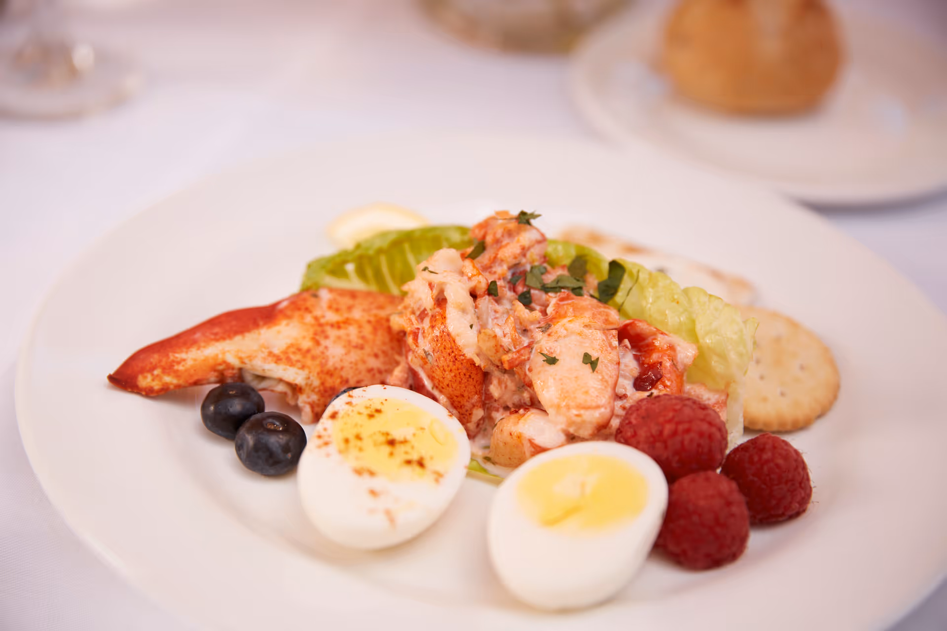 Plate with lobster salad, halved boiled eggs, raspberries, blueberries, and crackers on a white tablecloth.