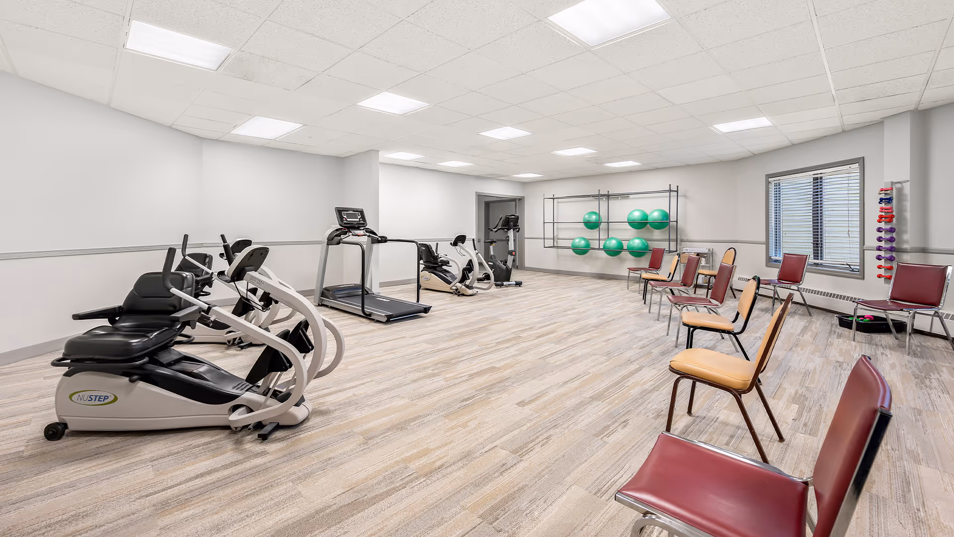 A spacious exercise room with light wood flooring and white walls. The room contains several pieces of exercise equipment including a recumbent bike, treadmill, and elliptical machines. There are several chairs arranged along the walls, and a rack holding green exercise balls and colorful dumbbells near a window.