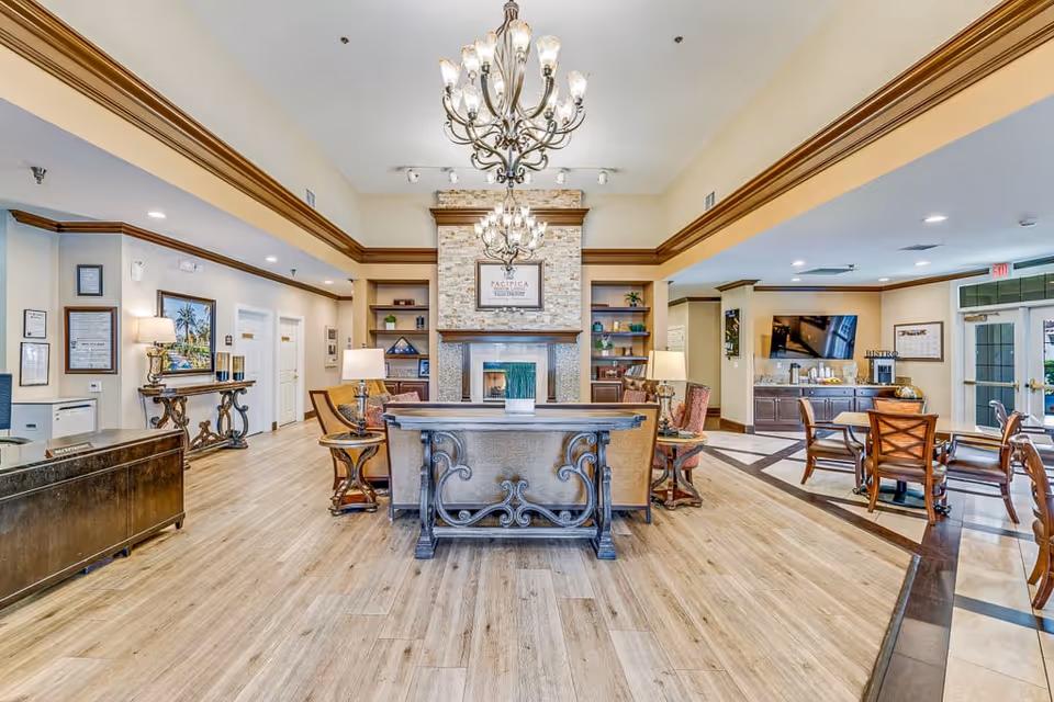 A spacious and well-lit senior living facility common area with wooden flooring, a decorative chandelier, a stone fireplace, comfortable seating, and a dining area with tables and chairs. The room features warm beige walls with crown molding and various framed pictures and decor items.