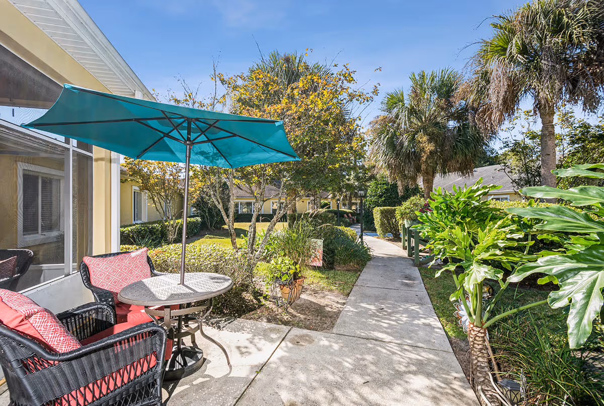 Outdoor patio area with a round table and two wicker chairs with red cushions under a teal umbrella. The patio is adjacent to a building with large windows, surrounded by lush greenery, palm trees, and a concrete walkway leading through the garden area under a clear blue sky.