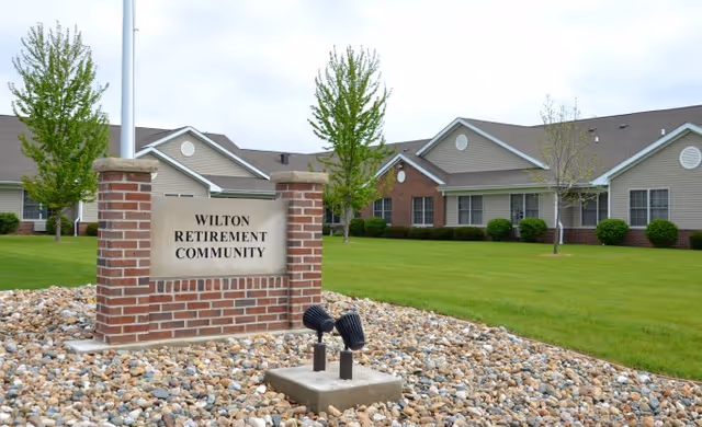 Exterior view of Wilton Retirement Community showing a brick sign with the community name in front of a well-maintained lawn and single-story residential buildings with beige siding and brick accents.