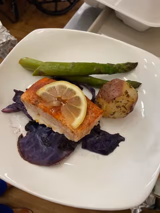 A white plate with a serving of cooked salmon topped with a lemon slice, two asparagus spears, a roasted potato, and some cooked purple cabbage.
