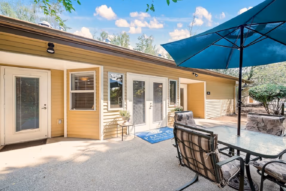 Outdoor patio area at The Cottages of Lynnwood featuring a glass-top table with cushioned chairs and a large blue umbrella. The patio is adjacent to a yellow building with multiple doors and windows, under a bright blue sky with scattered clouds.