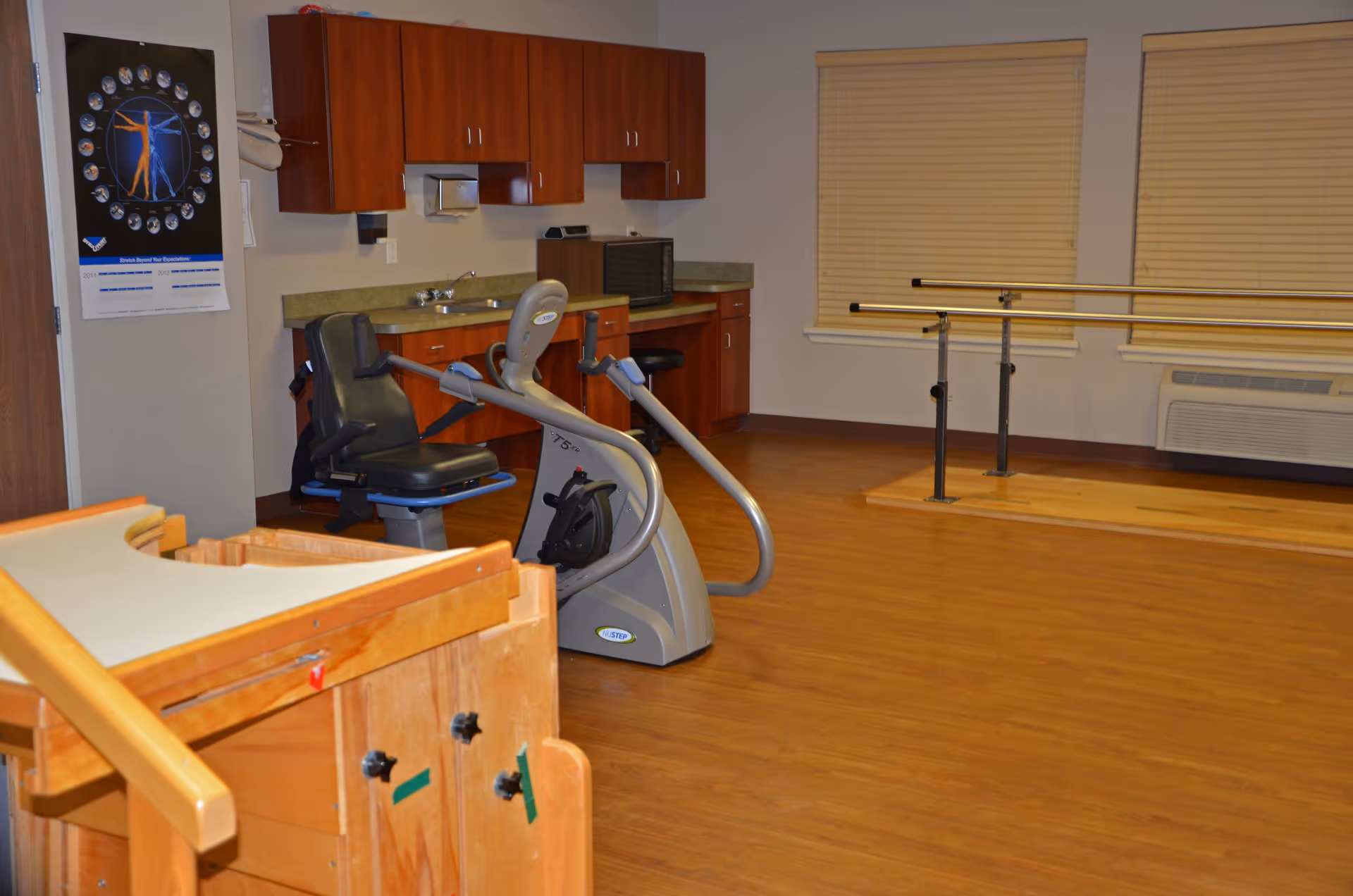A rehabilitation room with wooden flooring, a recumbent exercise bike, parallel bars for walking exercises, wooden therapy equipment, a countertop with cabinets, a microwave, and two windows with closed blinds.